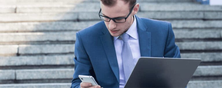 Geschäftsmann nutzt Smartphone und Laptop für mobiles Arbeiten, IT-Sicherheit im Fokus.